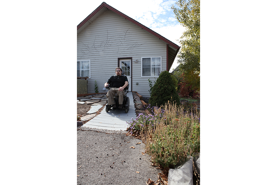 A green-shirted man travels in a power wheelchair down the wooden ramp toward the driveway.